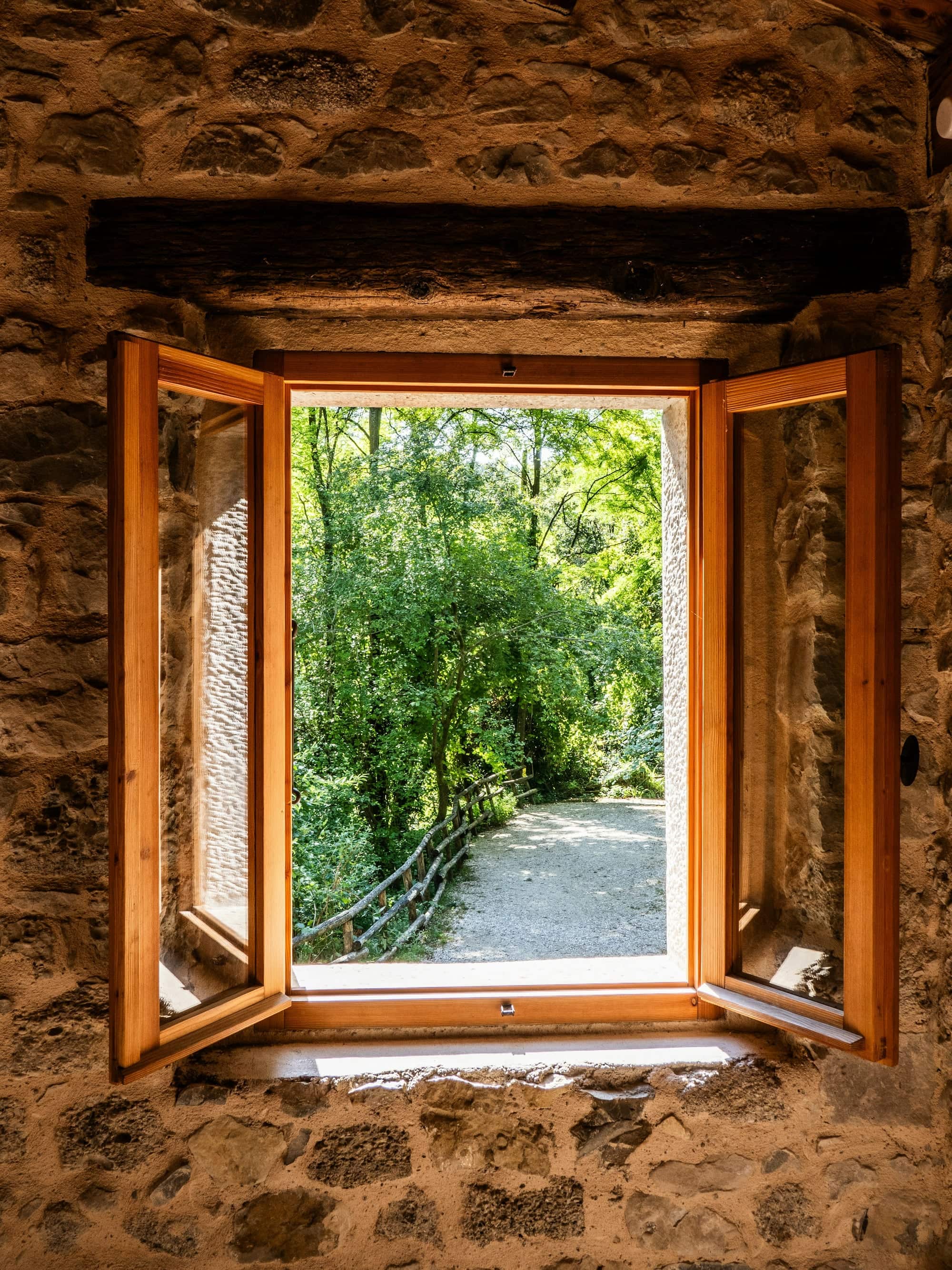 Fenêtre à deux battants en bois ouverte dans un épais mur de pierre, donnant sur un chemin bordé de verdure
